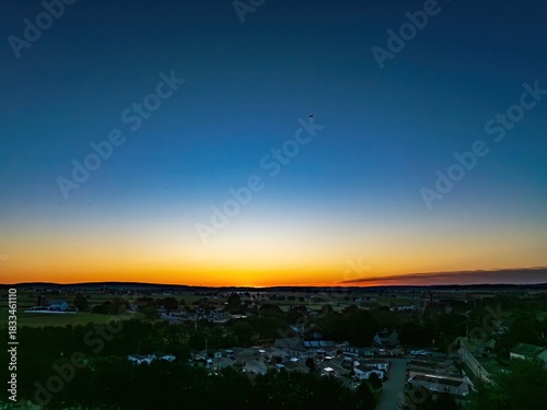 Wallpaper Mural A vibrant sunset casts warm colors across the sky above a small town. Fields stretch out in the distance, creating a serene atmosphere. The peaceful scene shows nature’s beauty as day turns to night. Torontodigital.ca
