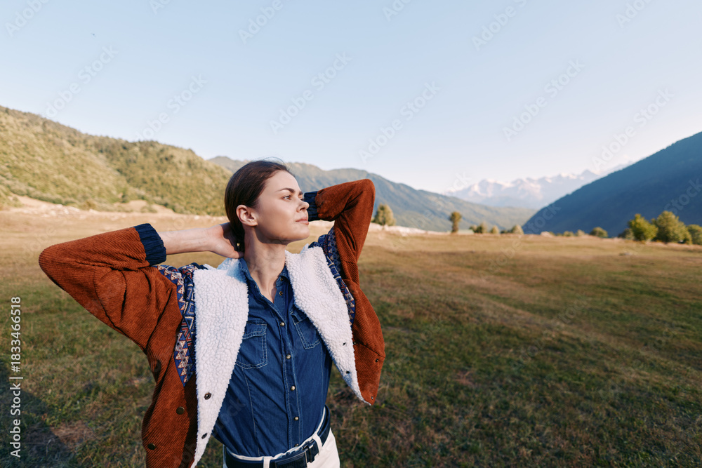 Fototapeta premium Woman outdoors mountains meadow relaxation pose, hands behind head, wearing jacket with sherpa and denim shirt; portrait of confident solo traveler enjoying scenic valley and clear sky.