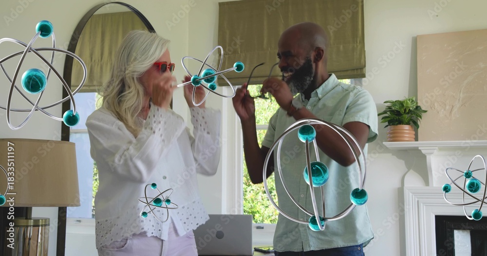 Naklejka premium Smiling senior couple interacting with floating atom models at home, with laptop and red sunglasses