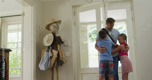 Hugging father wearing striped tee and messenger bag embracing kids at front door, with hat stand