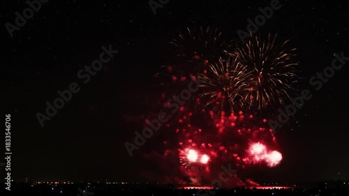 Bright red and white fireworks burst against a dark starry night sky over a distant city line