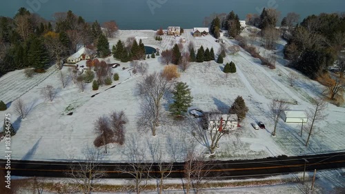 Aerial winter view of spacious residential lots with scattered houses, open snowy lawns, and mixed trees near a calm shoreline.