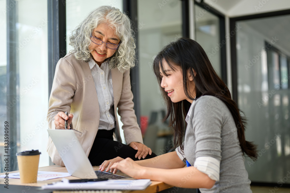 Fototapeta premium Old asian businesswoman sitting on table and talking to young coworker typing on laptop in office.