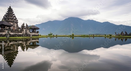 Scenic view of a Balinese temple reflected in a calm lake, with a mountain range in the background under a cloudy sky.
