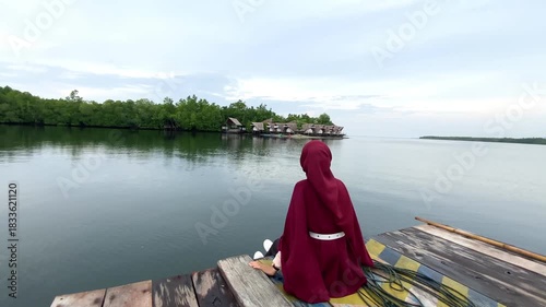 Young muslim woman wearing a maroon hijab is sitting calmly on a makeshift raft made by joining two boats together, with a wooden platform on top.
