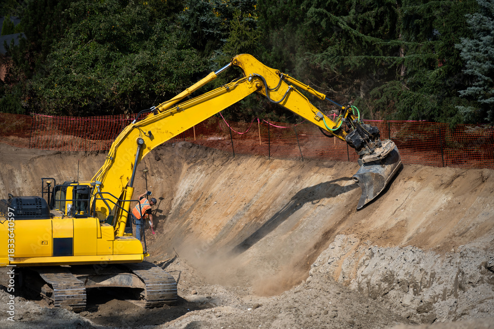Fototapeta premium Large earthworks project site with big excavator at bottom of foundation pit for a stormwater retention system, surveyor working with construction equipment operator