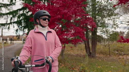 Woman standing with bicycle near vibrant red autumn trees, wearing helmet and sunglasses