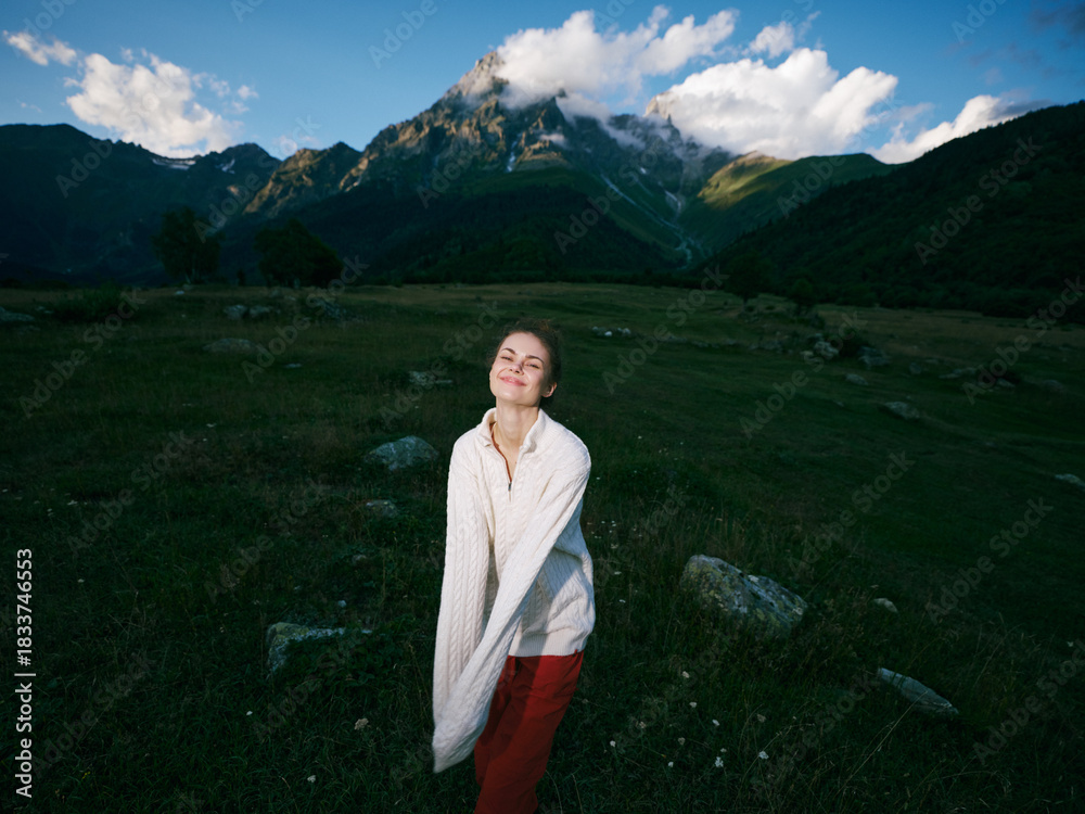 Obraz premium A woman standing in a grassy meadow with a dramatic mountain landscape, wrapped in a light blanket, enjoying fresh air and distant peaks in a peaceful outdoor moment