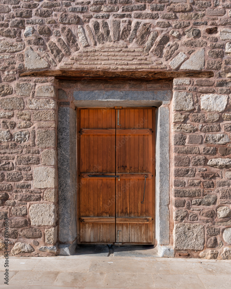 Fototapeta premium Double wooden door set in a textured stone wall with an arched stone lintel at Alaeddin Mosque, Konya, Turkey. Historic architecture detail