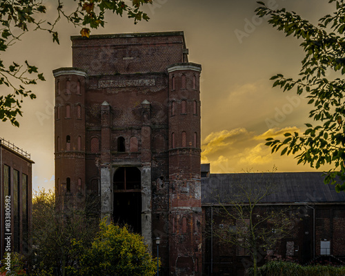 Das alte Zechengebäude, ein Riese aus Backstein, steht in der Dämmerung des Tages, während der Himmel dahinter in gedämpftem Gold und sanften Pastelltönen leuchtet. Die Fassade mit den vertikalen Böge