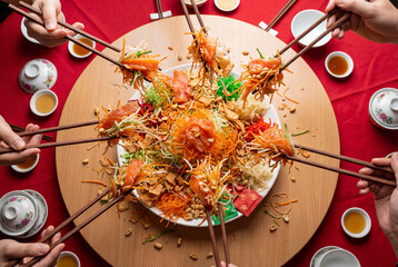 Multiple hands tossing salmon yusheng salad with chopsticks on a round table, festive shared meal for lunar new year celebration and family gathering tradition in Asian cuisine