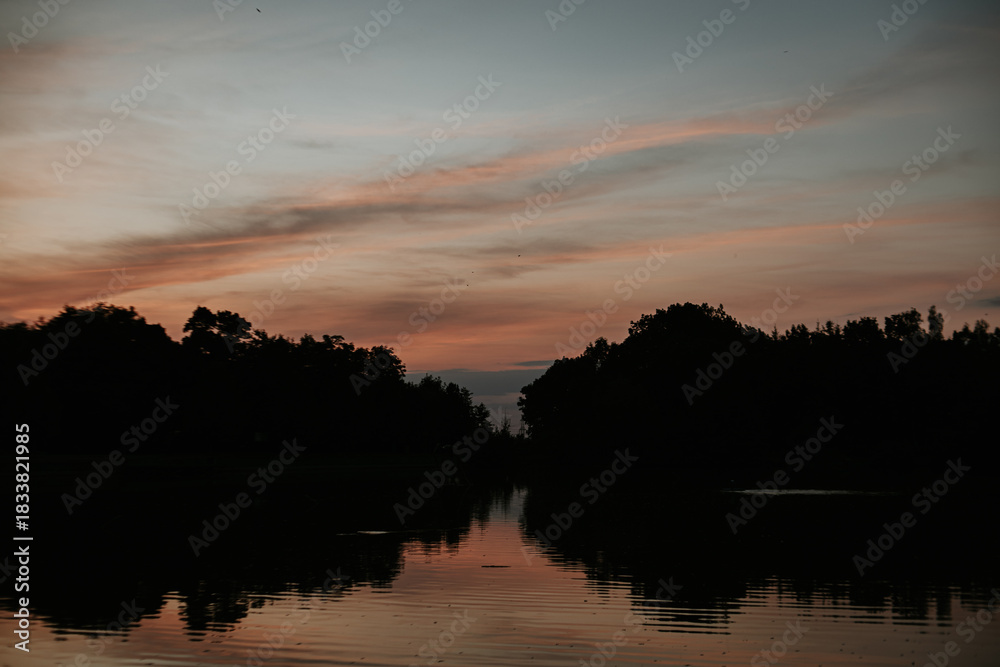 Fototapeta premium Sunset reflecting on calm lake water with dark forest silhouette