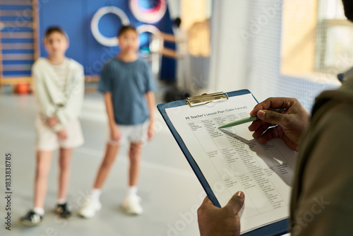 Two girls standing in gym while adult holding clipboard with lesson plan, focusing on physical education activity, kids waiting for instructions during school sports class