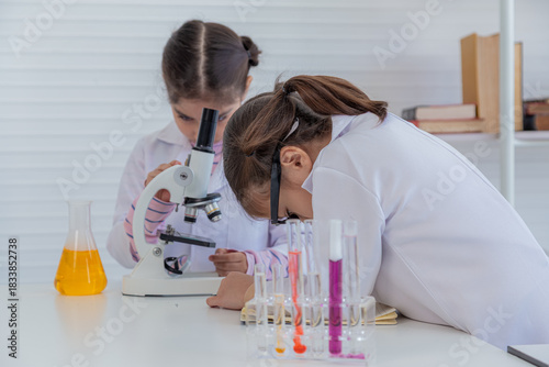 Adorable asian girls wearing lab coat whispering secrets and making experiment in chemistry class, little children whisper excitedly while science experiment together in classroom.