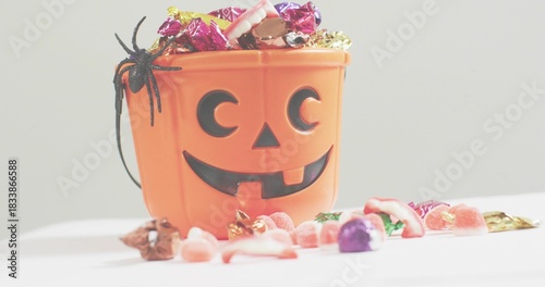 Showing jack o'lantern bucket holding foil candies and gummies on white table with black spider