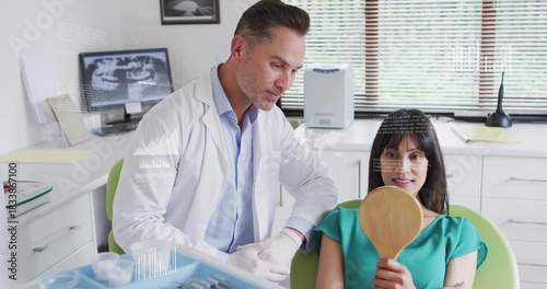 Leaning dentist in white coat gloved observing patient in green top at dental clinic holding mirror