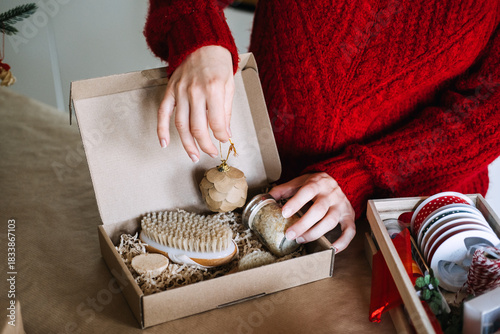 Person places ornament and jar of bath salts into cardboard gift box containing self-care items on table. Self-care gifting, plastic-free Christmas, eco packaging, sustainable lifestyle.