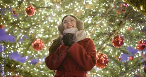 A young girl in a red coat walks in the evening at the European Christmas fair and admires her surroundings while standing against the backdrop of a Christmas tree with garlands and illuminations.