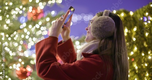 A young beautiful girl in a red coat walks in the evening at the European Christmas Market and takes pictures on a phone while standing against Christmas tree with garlands and illuminations.