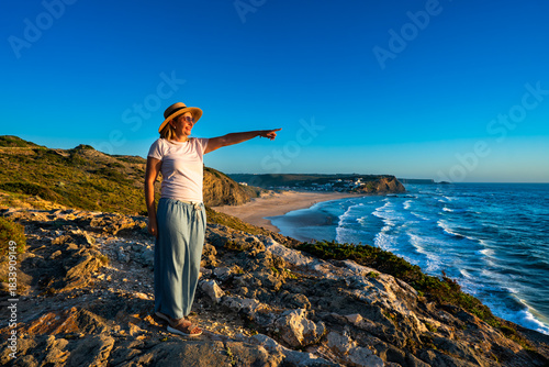 Beautiful middle-aged woman tourist standing on cliff overlooking Monte Clerigo beach and ocean in summer. Algarve coast in Portugal. Side view