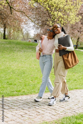 A female student hugs a female student walking next to her while they laugh and hold notebooks and a folder