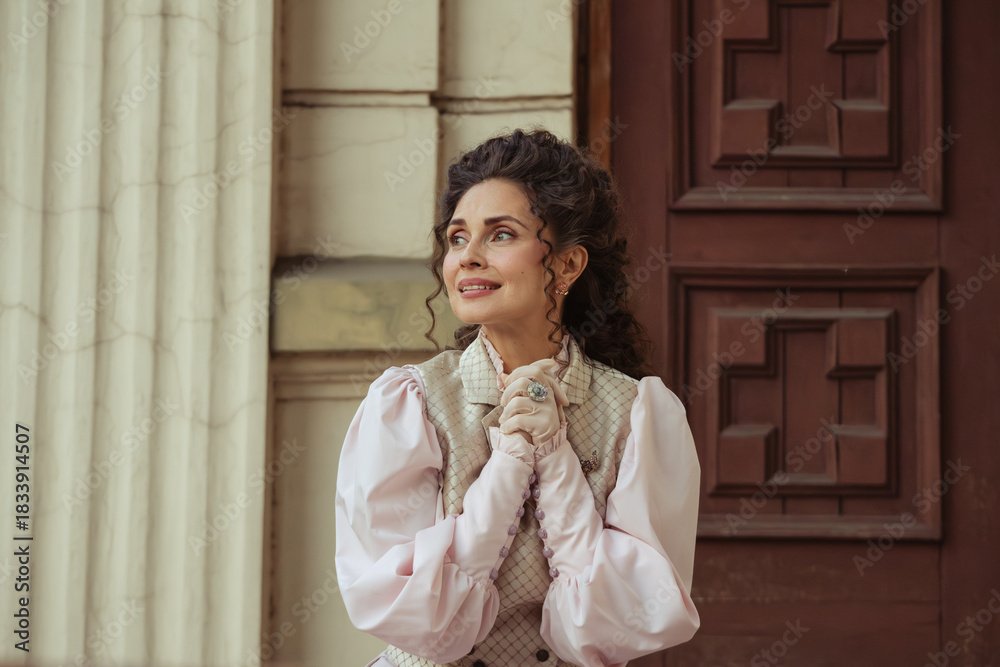 Fototapeta premium An elegant woman in a vintage dress sits next to an old stone column with a very happy expression on her face.