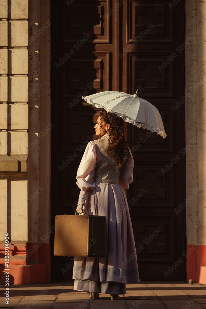 Fototapeta premium A woman in an antique dress with an umbrella and a suitcase stands in front of the doors of an old cathedral.