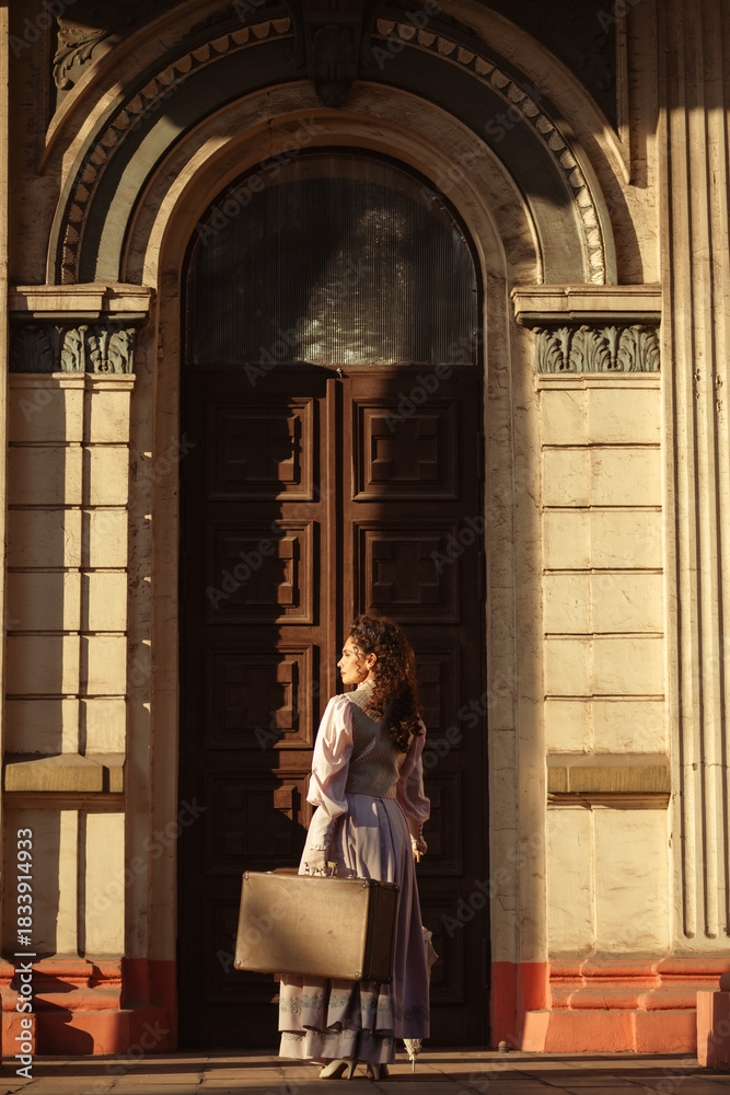 Obraz premium A woman in a vintage dress with a suitcase stands by ancient columns at dusk.