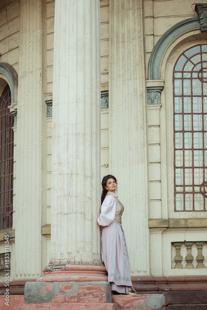Fototapeta premium An elegant woman in a vintage dress stands by an old stone column