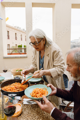 A woman stands and holds a spatula and fork in a frying pan while a man holds a plate and sits at a table while they talk