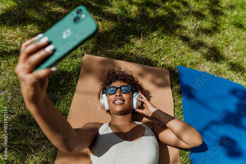 A woman looks at her phone and lies on a mat on the grass while listening to headphones