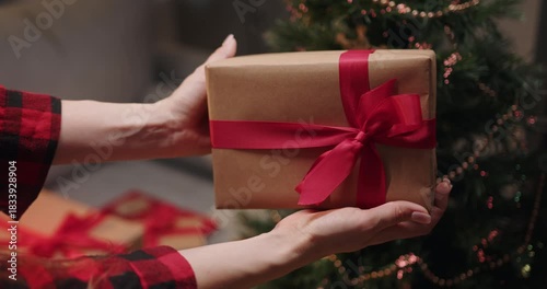 A person presents a wrapped gift with a red ribbon in front of a Christmas tree. A pregnant woman wearing a Santa hat opens a Christmas present in front of a decorated tree