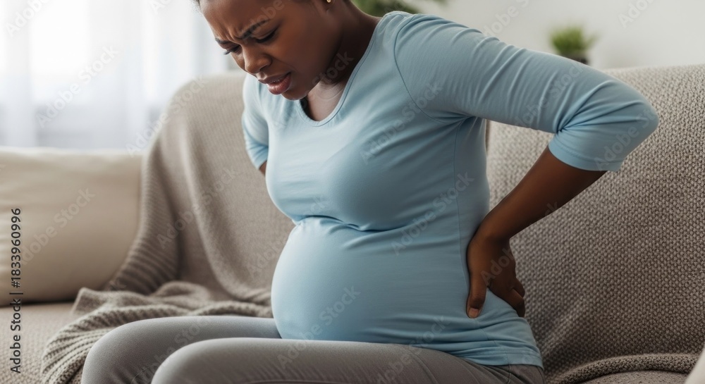 Obraz premium A pregnant woman suffering back pain, grimacing and holding her lower back while seated