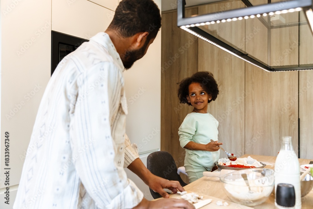 Obraz premium A daughter stands on a chair at the table and holds a spoon over a baking dish while listening to her father standing next to her