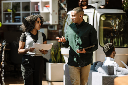 A male worker smiles and holds a computer tablet while talking to a female worker who walks next to him and listens while holding documents