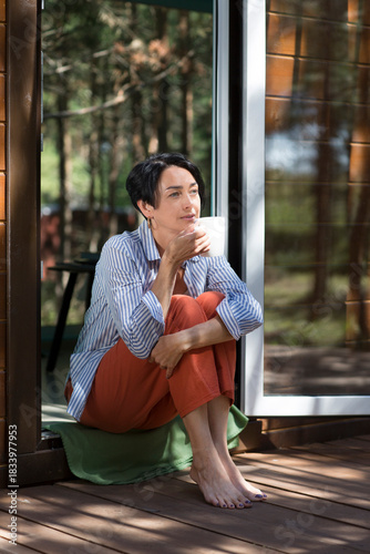Mindfulness and feel-good moments.  lifestyle portrait smiling middle aged woman drinking tea while sitting on the threshold in a country house in the forest. mental health. Enjoying the little things