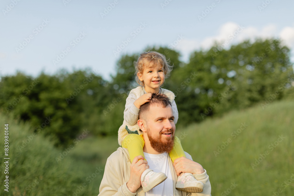Fototapeta premium Father with a red beard carrying his toddler on his shoulders while walking outdoors on a sunny day. Warm natural light, joyful family moment and peaceful countryside atmosphere.