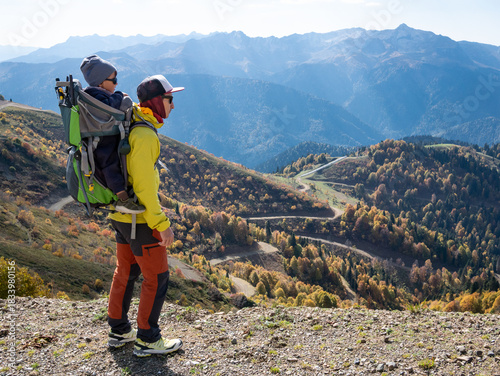 A man and his child in backpack carrier are hiking and looking at autumn mountains. Rosa Khutor resort in Krasnaya Polyana (Sochi) at sunny day. Not AI