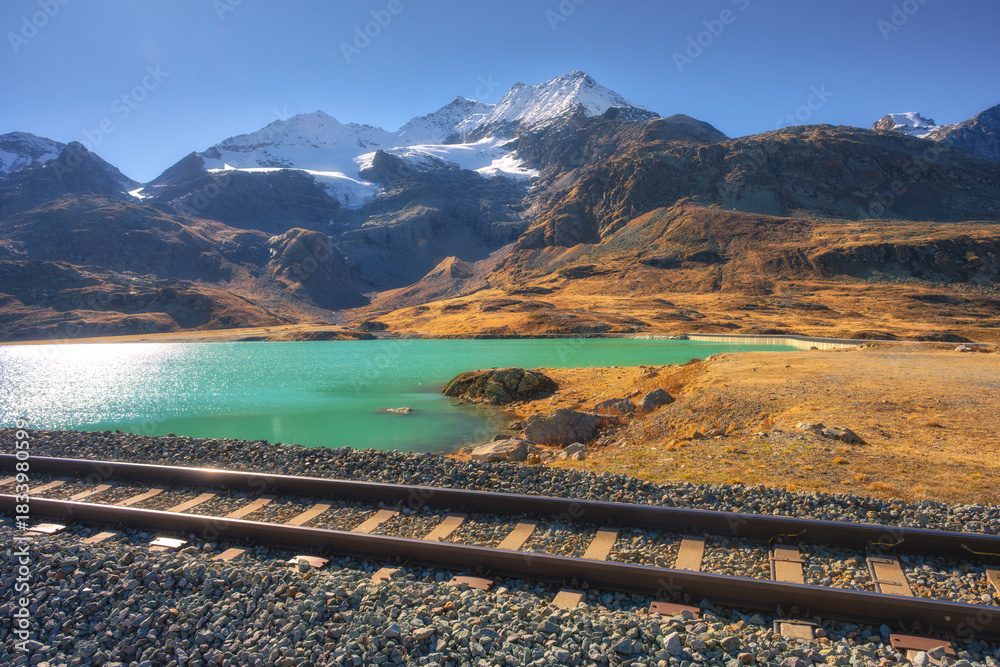 Obraz premium Scenic railway tracks running along turquoise lake Bianco with snowy alpine peaks on autumn sunny day. Swiss Alps landscape near Bernina Pass, train route in Switzerland. Railway station. Travel