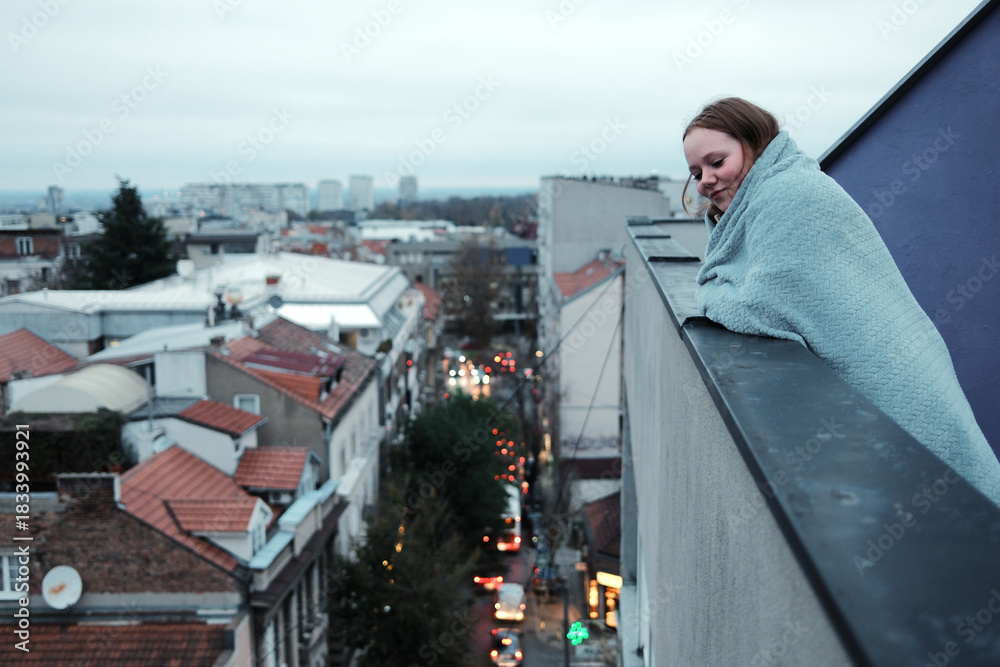 Obraz premium A woman wrapped in a blanket leans on a high balcony railing and observes the city street below. The concept highlights urban atmosphere and quiet reflection.