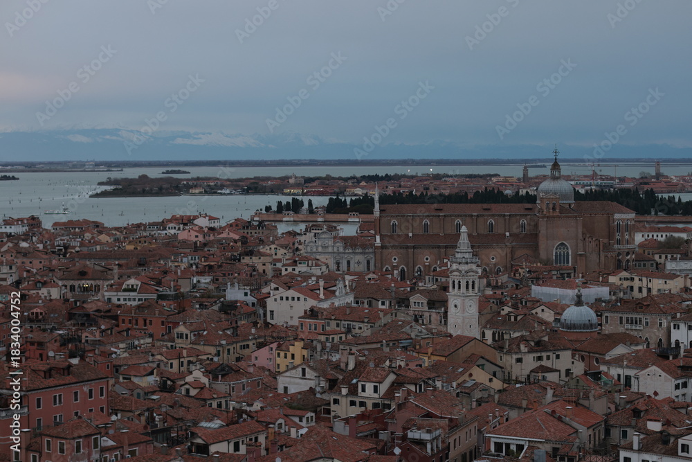Obraz premium Panoramic view of Venice, Italy, with terracotta rooftops, historic churches, and a large basilica near the lagoon under a cloudy evening sky.