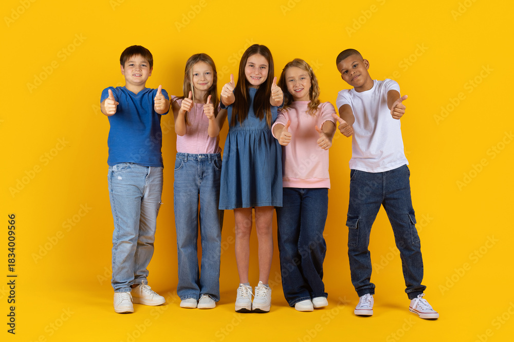 Obraz premium Preteen children standing in a row giving thumbs up in bright studio background. Confident and cheerful children expressing approval, unity and a positive school lifestyle mood