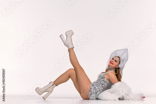 Woman in a silver sequin dress and kokoshnik lies on the floor, lifting one leg, with a white faux-fur coat on a clean studio background