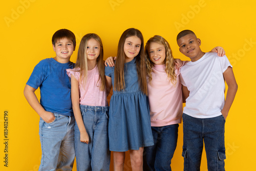 Smiling group of preteen kids embracing and posing on yellow studio background. Friendly children expressing closeness, confidence and a positive school lifestyle atmosphere