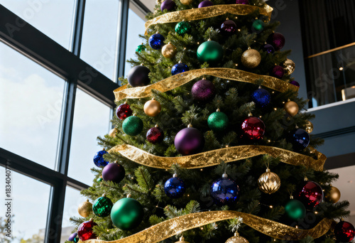 Decorated Christmas tree with colorful baubles and gold ribbon. Low angle view of festive pine in a modern interior with large windows. Holiday season background