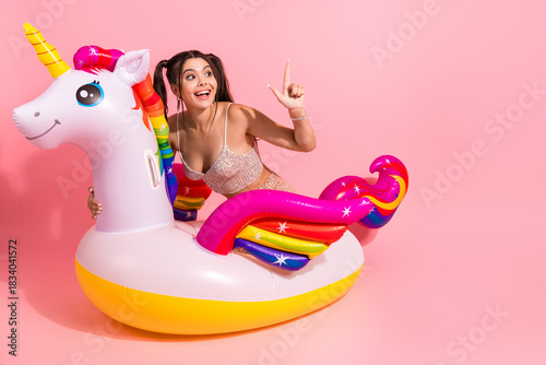 Young woman riding a unicorn inflatable float in a pink studio smiles and poses for fashion lifestyle and party promo