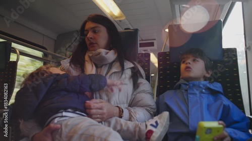 Mother sits on train holding restless toddler while older child looks upward creating authentic moment of overwhelmed parenting family travel fatigue and real everyday commute inside public transport