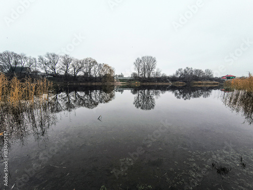 Canvas Print Tranquil lake with mirror-like reflections of leafless trees and cloudy sky, bordered by reeds and distant houses