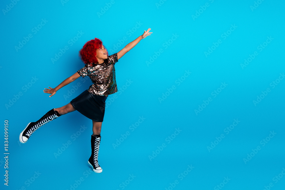 Naklejka premium Young girl with red hair wearing a shiny glitter top black skirt and high knee boots jumps joyfully against a bright blue background in a playful fashion moment