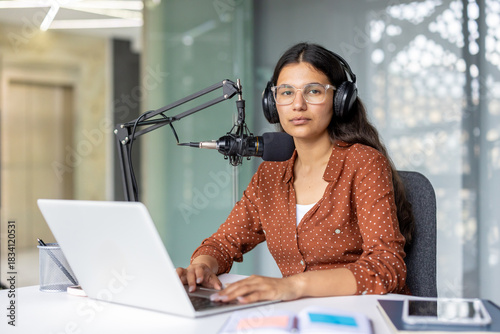 Young biracial woman with headphones and glasses sits at a desk with laptop and professional microphone, focused on recording or editing podcast and online audio content in home studio